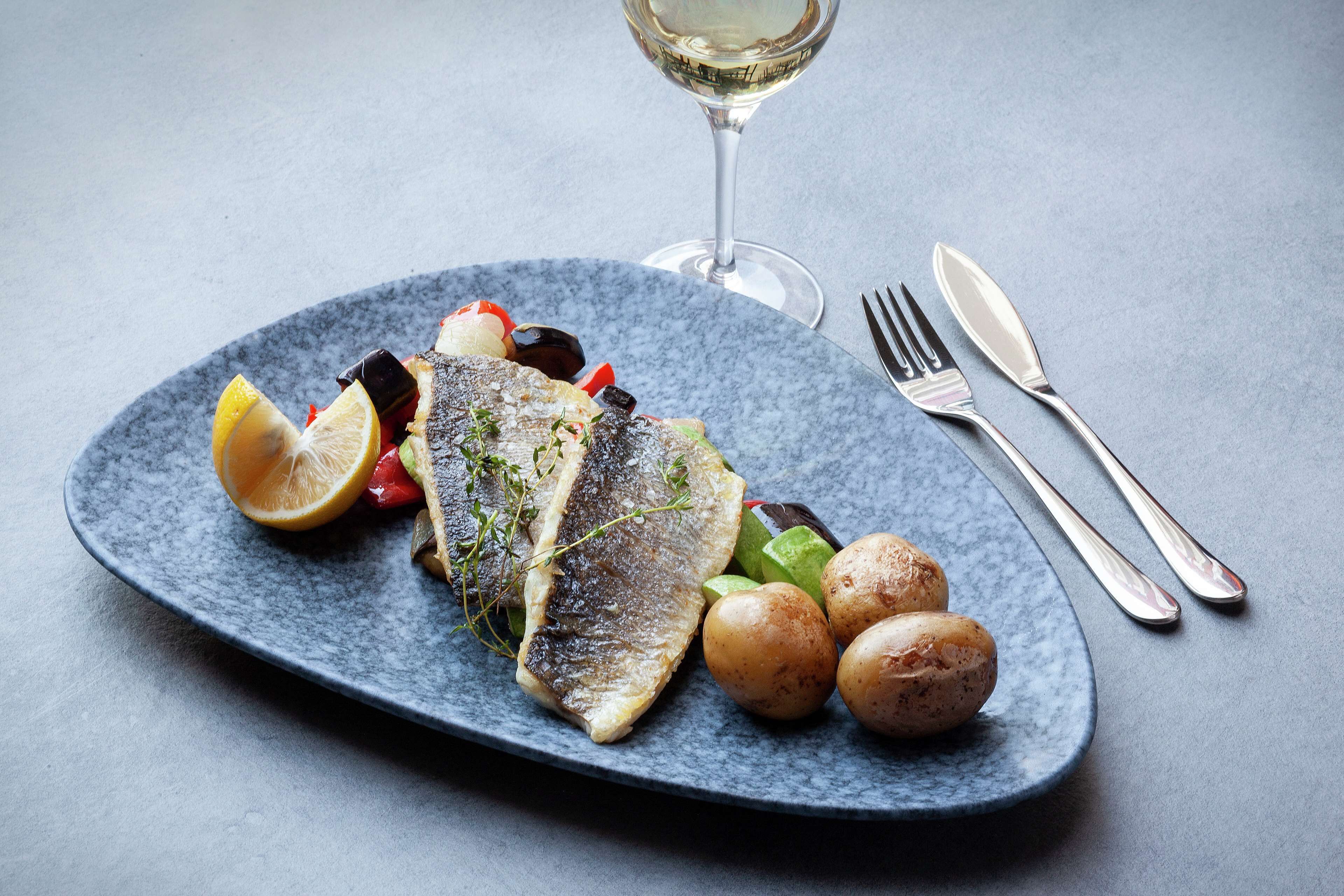 Ein Teller mit Fisch, Zitrone und Gemüse, dazu ein Glas Weißwein und Besteck auf einem blauen Teller.