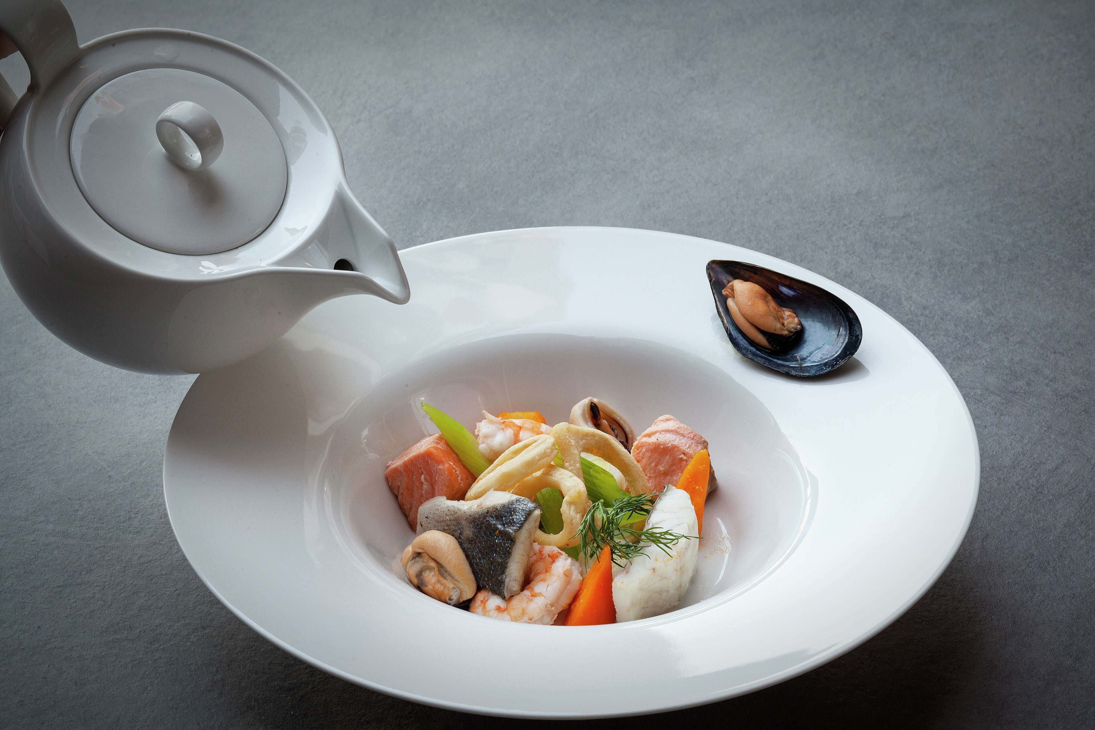 Une théière blanche verse du thé dans un bol blanc contenant divers fruits de mer et légumes.