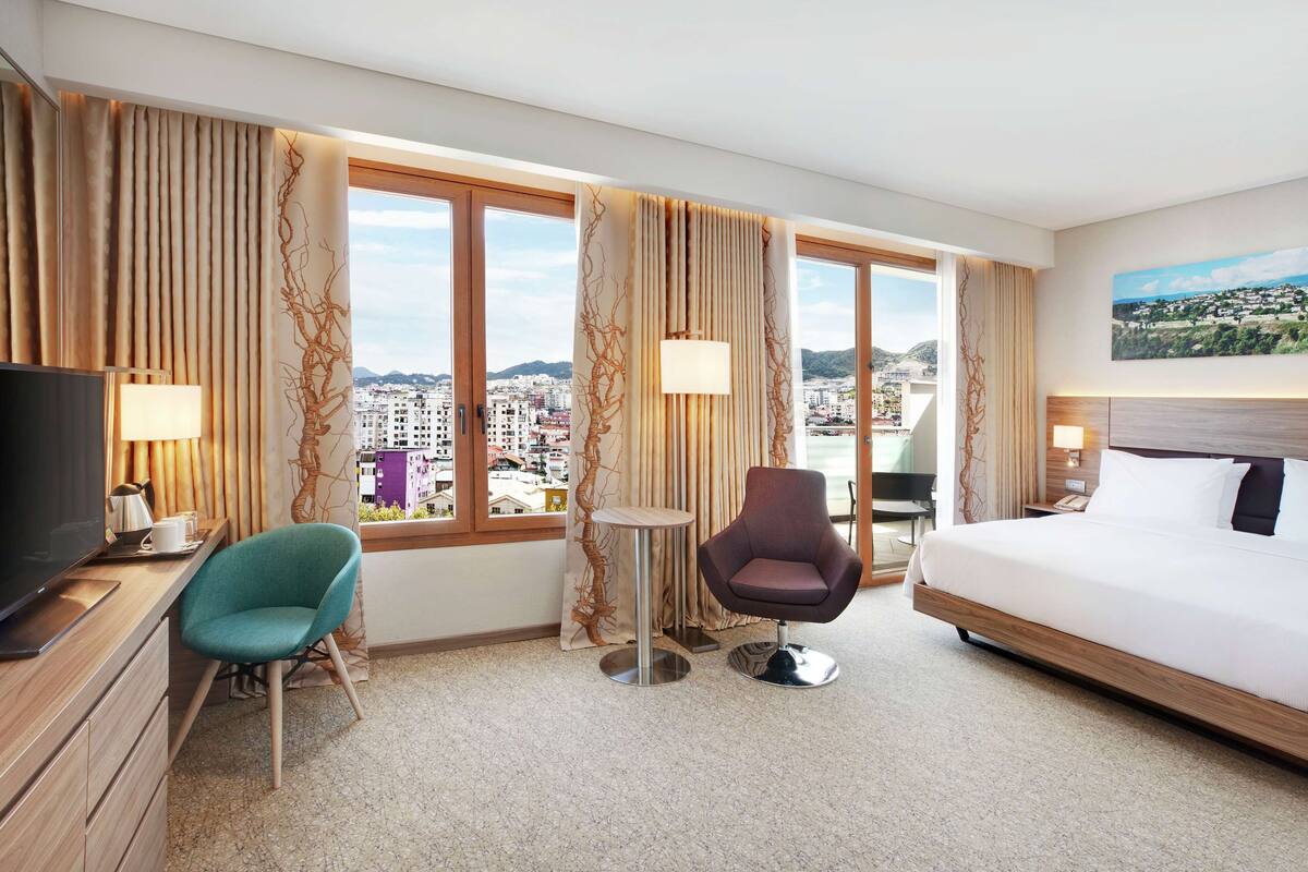 A hotel room with a bed, two chairs, a TV, and a view of a cityscape through large windows.