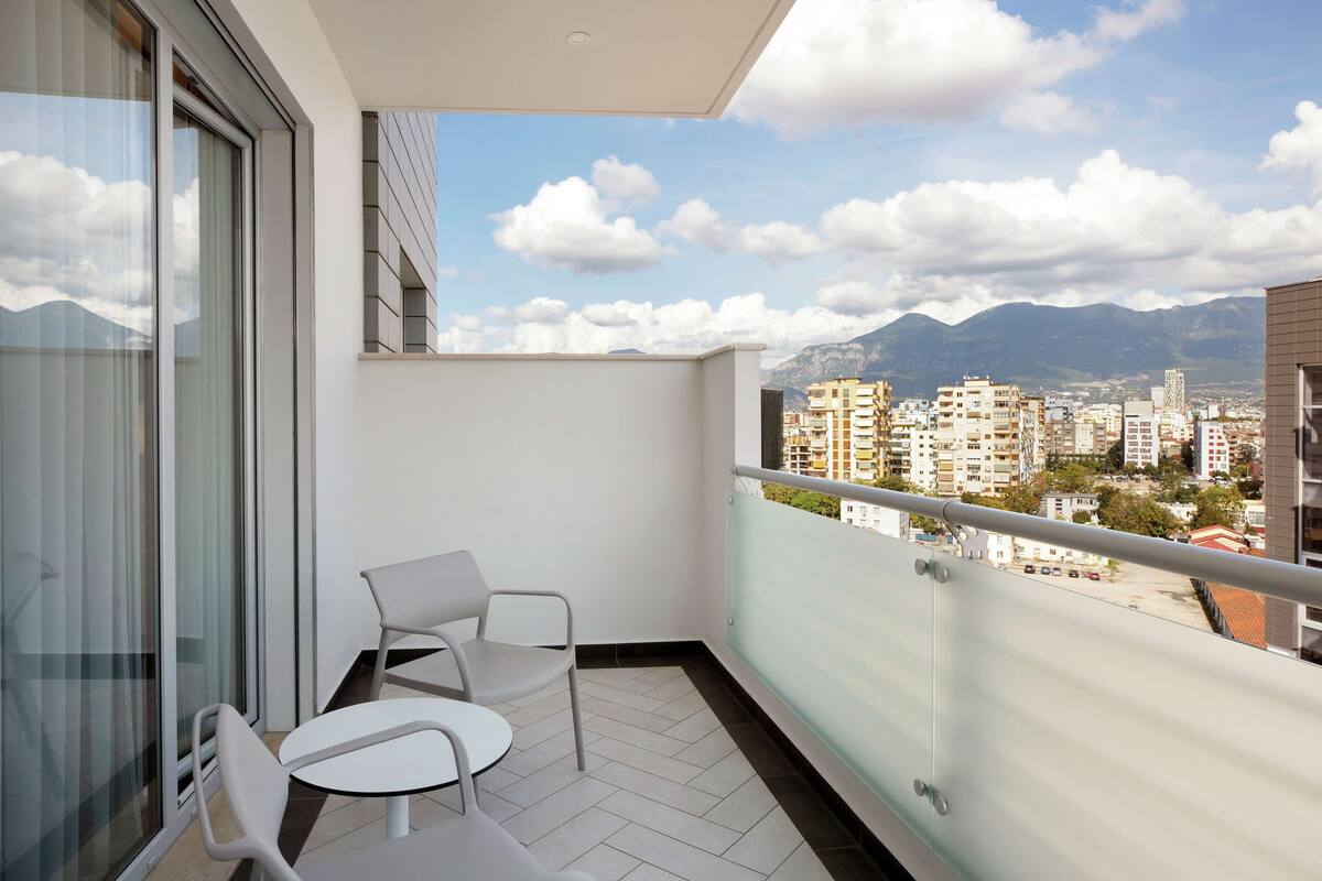 A balcony with two white chairs and a table, overlooking a cityscape with mountains in the background.