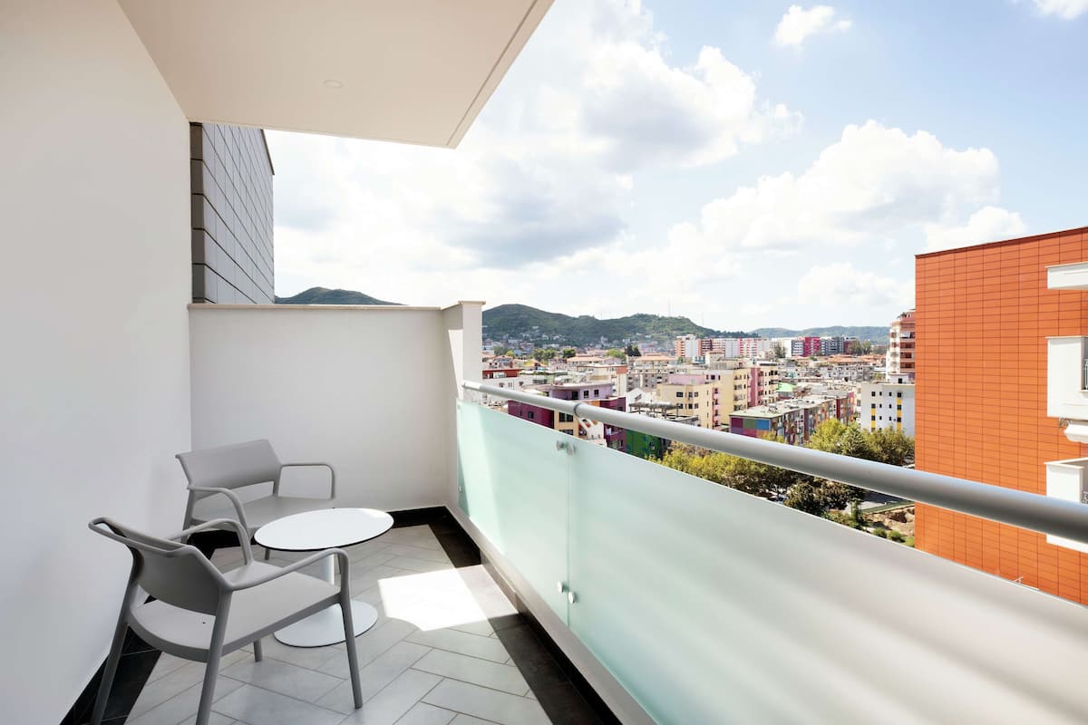 A balcony with two chairs and a small table, offering a view of a cityscape with buildings and hills in the background.