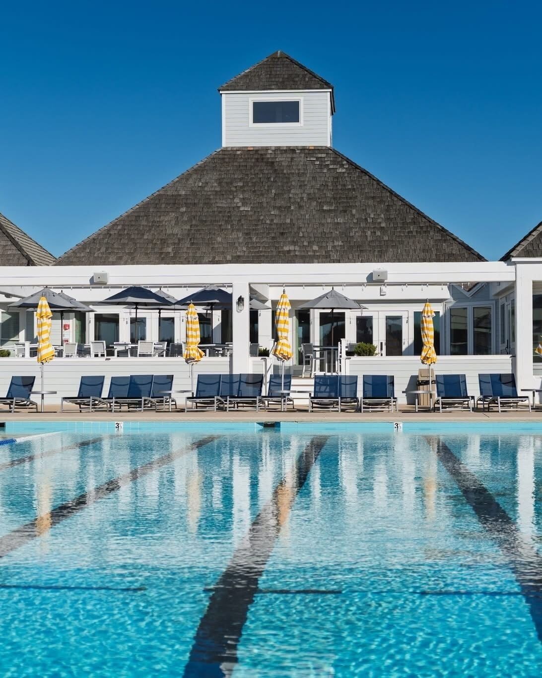 A swimming pool with lounge chairs and umbrellas, a covered seating area, and a building with a gabled roof.