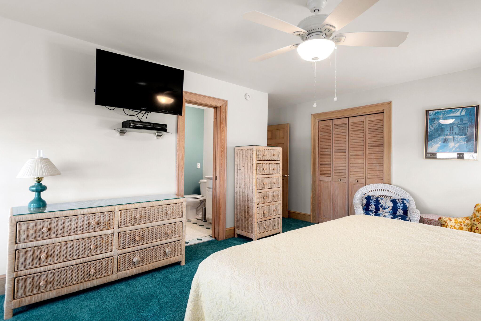 Un dormitorio con cama, cómoda, televisor montado en la pared, ventilador de techo y puerta que da al baño.
