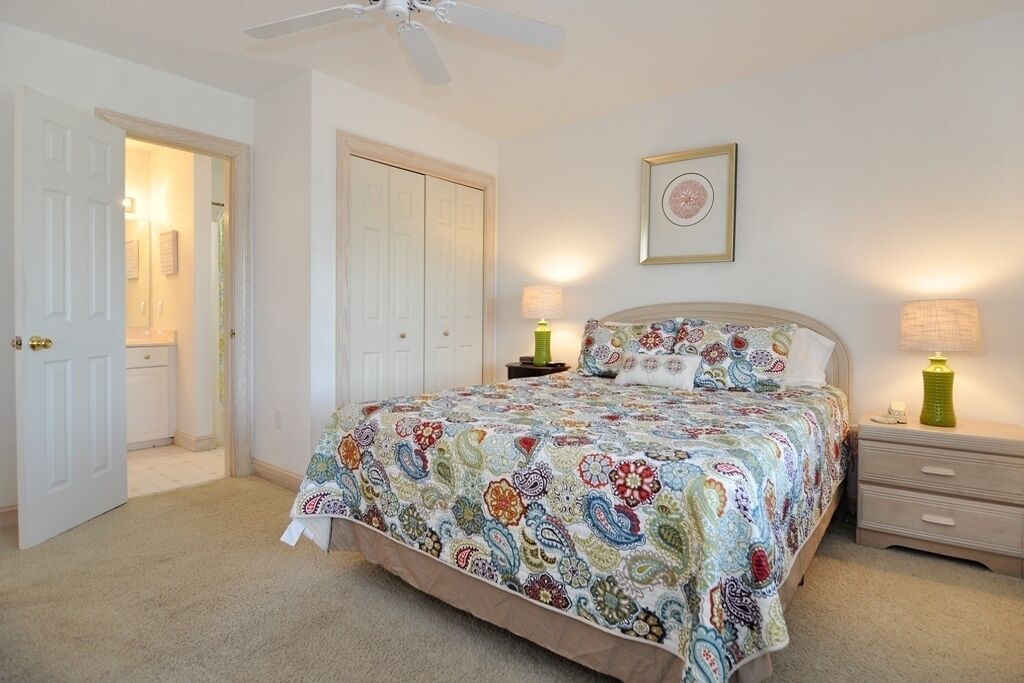 A bedroom with a bed, bedside lamps, a ceiling fan, and a door leading to another room.