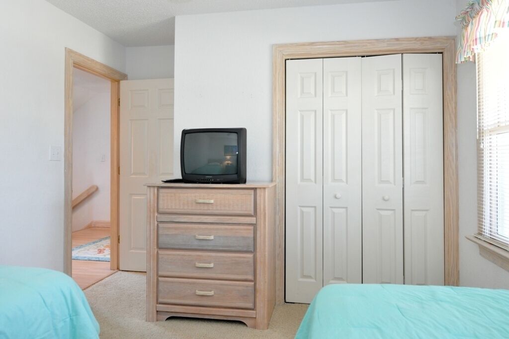 A bedroom with a wooden dresser, a television on top, a white door, and a window with blinds.