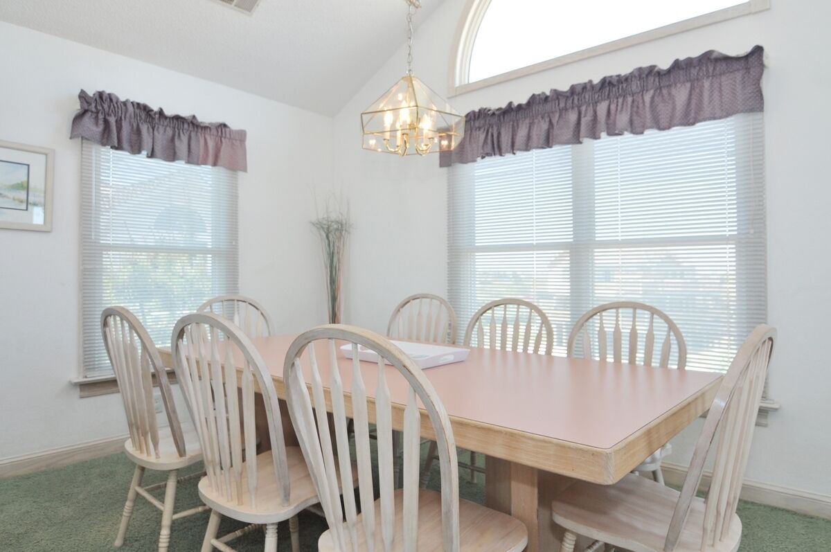 A dining room with a wooden table and six chairs, large windows with blinds, a chandelier, and a framed picture on the wall.