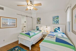 A bedroom with two beds, a ceiling fan, artwork on the walls, and a window with blinds.
