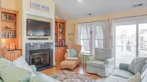 A cozy living room with a fireplace, bookshelves, a television, and a view of the city. - Heels In The Sand: Close to beach, Private Pool (Nags Head)