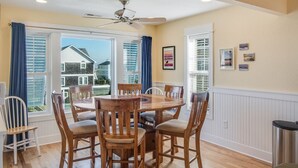 A dining area with a round wooden table and chairs, large windows with blinds, a ceiling fan, and a view of neighboring houses. - Heels In The Sand: Close to beach, Private Pool (Nags Head)