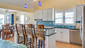 A kitchen with white cabinets, a blue hanging light, and a dining area with wooden chairs. - Heels In The Sand: Close to beach, Private Pool (Nags Head)