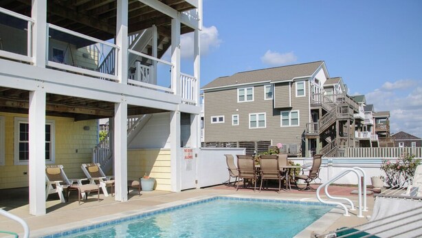 A pool area with lounge chairs and a dining table, a multi-story building with balconies, and a clear sky. - Heels In The Sand: Close to beach, Private Pool (Nags Head)