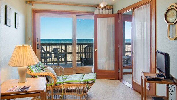 A room with a wicker sofa, a table with a lamp, a television, and a view of the ocean through a sliding glass door. - Heron Cove 202S: Oceanfront Condo, Shared Pool (Nags Head)