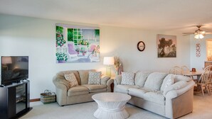 A living room with two sofas, a coffee table, a TV, and artwork on the walls. - Heron Cove 202S: Oceanfront Condo, Shared Pool (Nags Head)