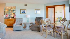 A living room with a beige sofa, two armchairs, a glass coffee table, and a dining table with wicker chairs. There are framed pictures on the wall and a cabinet with decorative items. - Heron Cove 202S: Oceanfront Condo, Shared Pool (Nags Head)