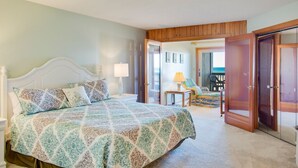 A bedroom with a large bed, a nightstand, a chair, and a door leading to another room. - Heron Cove 202S: Oceanfront Condo, Shared Pool (Nags Head)