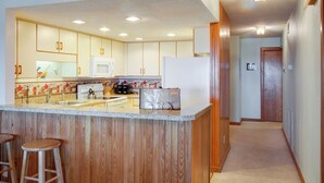 A modern kitchen with wooden cabinets, a central island with stools, and a microwave. - Heron Cove 202S: Oceanfront Condo, Shared Pool (Nags Head)