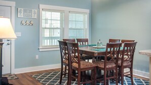 A dining area with a table and chairs, a lamp, and a window with blinds. - Star of the Sea: Oceanfront Condo, Shared Pool (Kill Devil Hills)