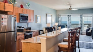 A modern kitchen with a central island, stainless steel appliances, and a view to the outside. - Star of the Sea: Oceanfront Condo, Shared Pool (Kill Devil Hills)