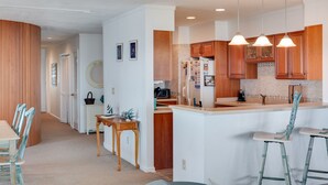 A modern kitchen with wooden cabinets, a central island, pendant lighting, and a dining area with a table and chairs. - Daniel's Diamond: Oceanfront, Great Views, Pool (Nags Head)