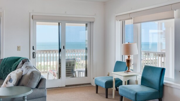 A room with a view of the ocean, a balcony with a railing, a table with a lamp, and a blue chair. - Daniel's Diamond: Oceanfront, Great Views, Pool (Nags Head)