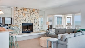 A living room with a stone fireplace, a grey sofa, a round coffee table, and a TV on a stand. - Daniel's Diamond: Oceanfront, Great Views, Pool (Nags Head)