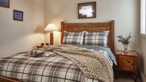 Ein Schlafzimmer mit kariertem Überwurf, einem Holzkopfteil, einem Nachttisch mit Lampe und einer kleinen Pflanze.