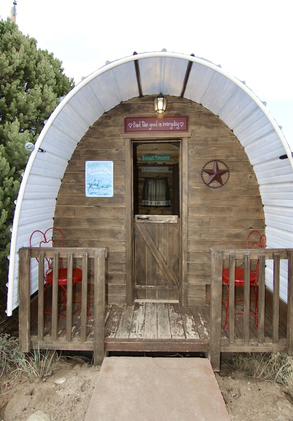 A rustic wooden cabin with a curved metal roof, a wooden door, and a small porch with red chairs.