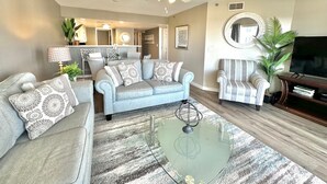 A living room with a sofa, armchairs, a coffee table, and a TV. There is a ceiling fan, a dining area in the background, and a plant on the right. - Summer & July 4th Savings at Beachside Colony Tybee's ONLY Oceanfront Resort 2/2 (Tybee Island)