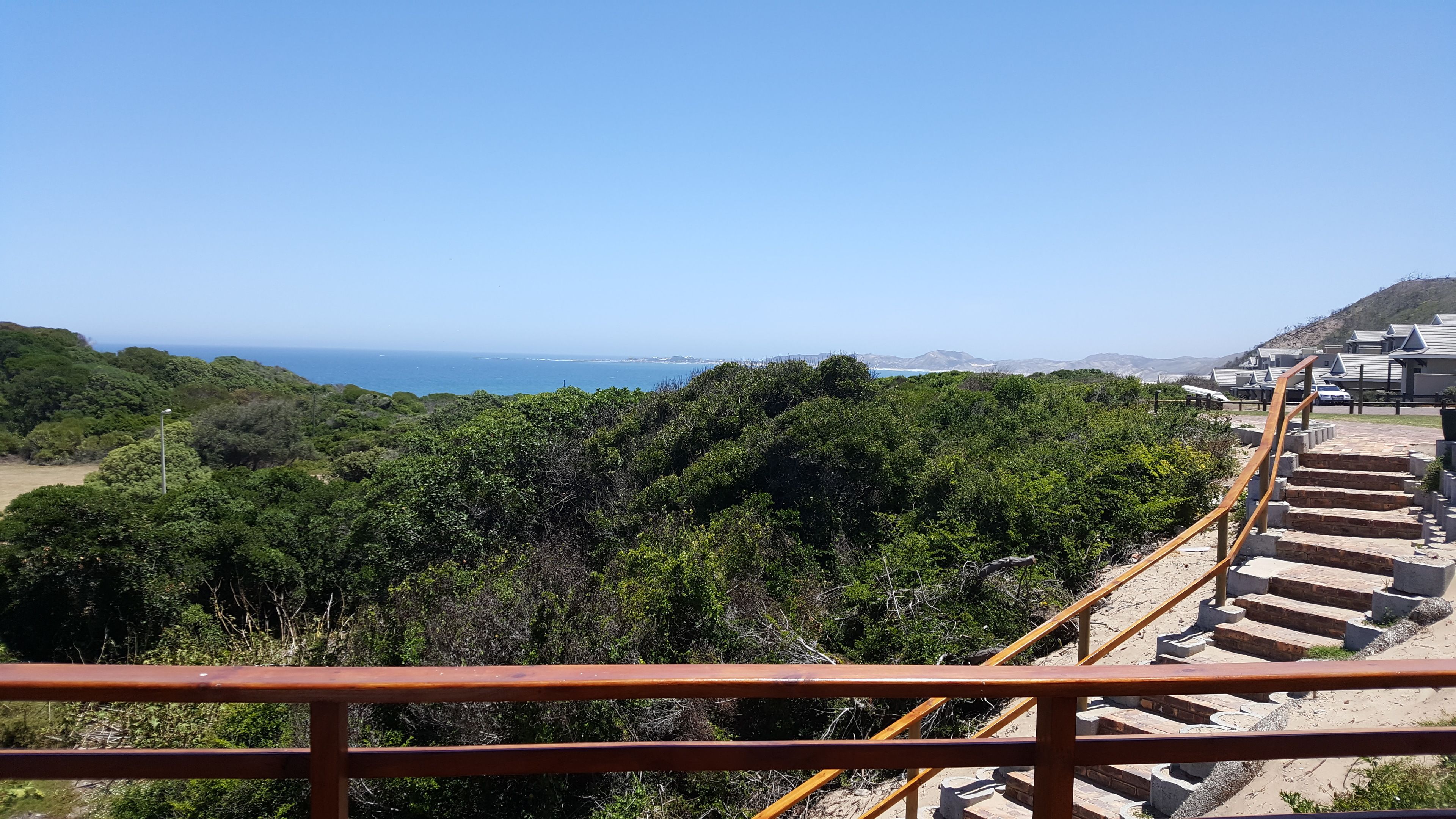 A coastal view with a staircase leading down to a sandy area, surrounded by dense greenery and overlooking the sea.