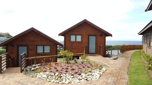 Two wooden cabins with a paved pathway leading to a garden area with rocks and flowering plants. - Brenton On Sea Cottages (Knysna)