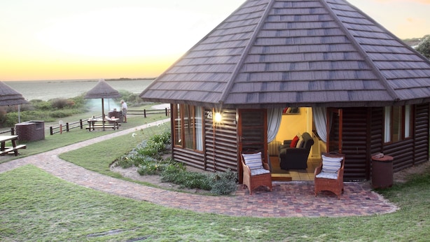 A lodge with a conical roof, a patio with wicker chairs, and a view of the sea. - Brenton On Sea Cottages (Knysna)