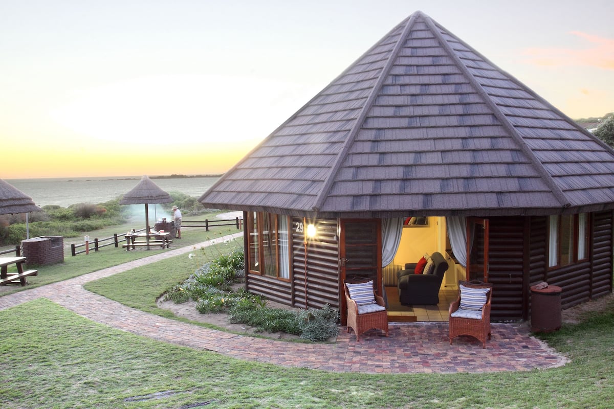 Eine Lodge mit einem kegelförmigen Dach, einer Terrasse mit Korbstühlen und Blick auf das Meer.