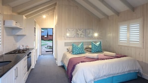 A bedroom with a bed, a nightstand, a built-in desk, and a window with blinds. - Brenton On Sea Cottages (Knysna)