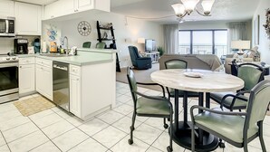 A modern kitchen with white cabinets, a dining area with a round table, and a living space with a sofa and TV. - Chateaux 403-Direct Gulf Front Condominium (Indian Shores)