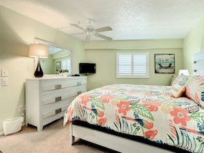 A bedroom with a bed, dresser, ceiling fan, TV, and a window with blinds.
