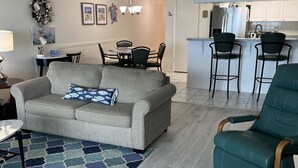 A living room with a grey sofa, teal armchair, and patterned rug. A dining area with chairs and a table is visible in the background. - Chateaux 403-Direct Gulf Front Condominium (Indian Shores)