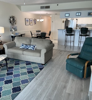 A living room with a grey sofa, teal armchair, and patterned rug. A dining area with chairs and a table is visible in the background.