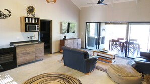 A modern living room with a sofa, ottoman, and a dining area with chairs. There is a kitchen area with a microwave and a clock. - Wild Dog Guest Lodge (Hoedspruit)