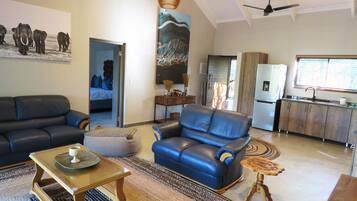 A modern living room with a sofa, armchair, and coffee table. There is a kitchen area with a refrigerator and a window.