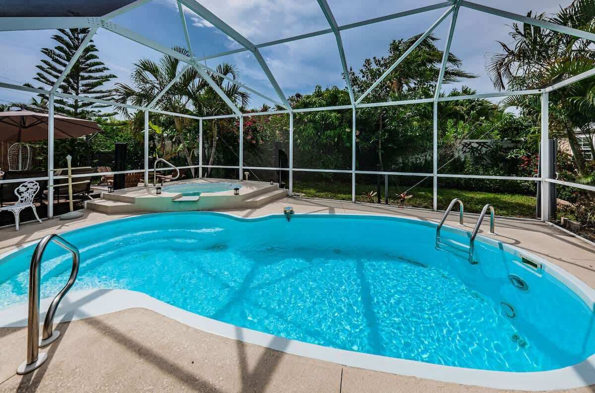 Una piscina con cerramiento de vidrio, rodeada de palmeras y un patio con sillas y sombrillas.