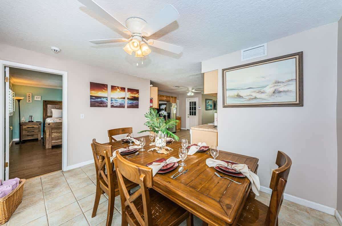 Un coin repas avec une table en bois dressée pour six personnes, un ventilateur de plafond et un tableau accroché au mur.