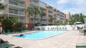 A hotel pool area with multiple lounge chairs, a swimming pool, and a multi-story building with balconies.