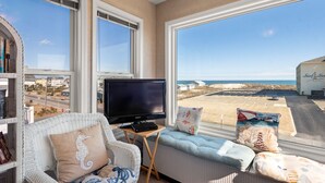 A room with a large window offering a view of the beach, a television on a small table, and a wicker chair with a cushion. - No Regrets: Semi-Oceanfront, Private Pool, 2 Spas (Nags Head)