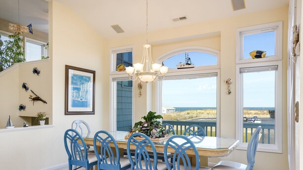 A dining area with a table and chairs, a chandelier, and large windows with a view of the beach. - No Regrets: Semi-Oceanfront, Private Pool, 2 Spas (Nags Head)