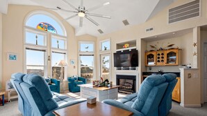 A spacious living room with a fireplace, large windows, and a ceiling fan. - No Regrets: Semi-Oceanfront, Private Pool, 2 Spas (Nags Head)