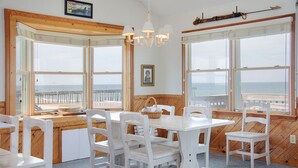 Un espace repas avec des meubles en bois blanc, de grandes fenêtres offrant une vue sur l’océan et un plafond en bois.