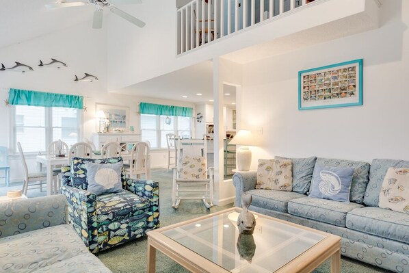 A spacious living room with a high ceiling, a glass coffee table, a blue sofa, a patterned armchair, and a dining area with white chairs.