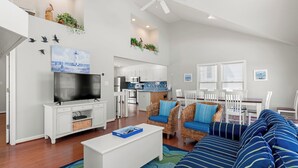 Ein modernes Wohnzimmer mit Flachbildfernseher, weißen Möbeln, einem blau gestreiften Sofa und einer Küche im Hintergrund.