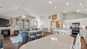A spacious living area with a large dining table, a fireplace, and a kitchen with white cabinets and modern appliances. - Sandy Sea Gulls: Private Hot Tub, Short walk to Beach & Community Pool (Nags Head)