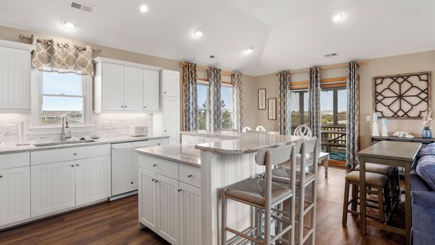A modern kitchen with white cabinetry, a central island, and a dining area with a wooden table and chairs. - Sandy Sea Gulls: Private Hot Tub, Short walk to Beach & Community Pool (Nags Head)